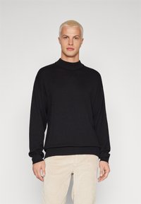 Sudadera negra de punto con cuello alto, mangas largas y puños acanalados. Combinada con pantalones de pana de color claro.