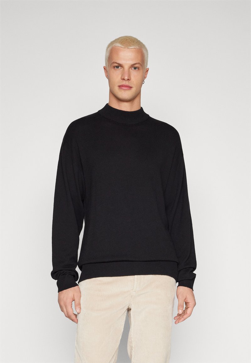 Sudadera negra de punto con cuello alto, mangas largas y puños acanalados. Combinada con pantalones de pana de color claro.