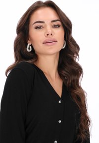 Fekete kardigán V-nyakkivágással, díszes gombokkal és puha textúrával. A modell csillogó fülbevalót visel, hosszú, hullámos haj keretezi az arcát.