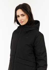 Chaqueta negra aislante con cierre de cremallera, capucha con reguladores ajustables y dos bolsillos frontales. Textura suave con sutil branding.