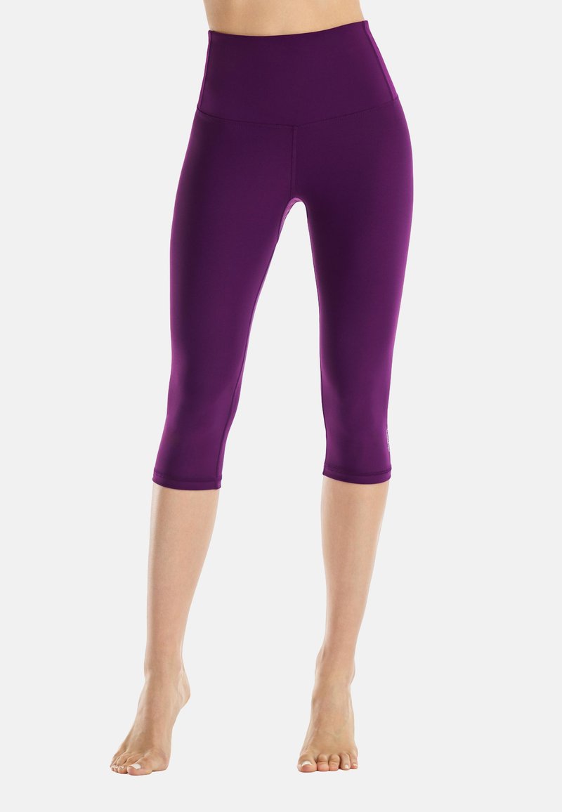 Gambe con leggings sportivi viola, a vita alta e taglio corto, in piedi a piedi nudi su sfondo bianco.