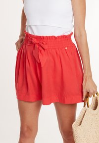 Koraalrode shorts met een smocktailleband en strikdetail. Lichte, vloeiende stof met plooien, gecombineerd met een geweven tas.