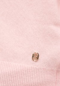 Ανοιχτό ροζ πλεκτό ύφασμα με ριμπ τελείωμα και μικρό λαμπερό χρυσό οβάλ έμβλημα που φέρει αφηρημένο, δεμένο σχέδιο.