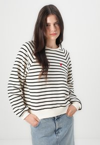 Stripet genser i krem med svarte striper, med en liten rød hjerte detalj på brystet. Laget av et mykt og behagelig stoff.