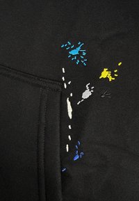 Zwarte stof met een gestikte zak, versierd met kleine geborduurde verfspetters in blauw, geel, wit en zwarte draad.