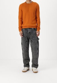 Pomarańczowy prążkowany sweter z dzianiny w połączeniu z czarnymi dżinsowymi spodniami typu cargo z przeszyciami i wieloma kieszeniami. Obuwie: białe trampki.