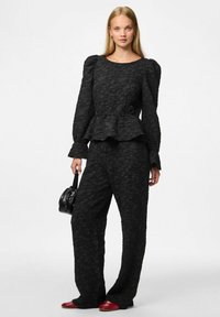Czarna, teksturowana bluzka z peplum i bufkami, zestawiona z luźnymi spodniami; modelka trzyma małą czarną torbę i ma na sobie czerwone buty.