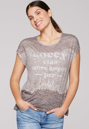 Hellgraues T-Shirt mit strukturierter Oberfläche, verziert mit metallisch silberner Schrift. Kurze Ärmel und eine lässige Passform. Getragen mit blauen Jeans.