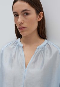 Top in tessuto morbido di un azzurro chiaro con scollo rotondo, dettagli arricciati e bordo a contrasto di un azzurro chiaro, che mette in evidenza una vestibilità rilassata e una texture liscia.