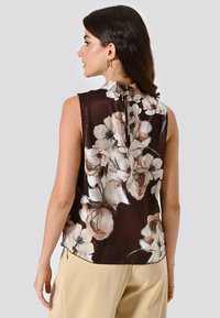 Donna vista di spalle con blusa senza maniche a fiori scuri e pantaloni beige, con capelli castani di lunghezza media e orecchini a cerchio.