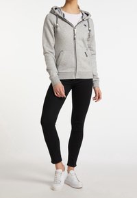 Sudadera gris con cremallera y capucha ajustable, dos bolsillos frontales y puños de canalé. Combinada con jeans negros ajustados y zapatillas blancas.