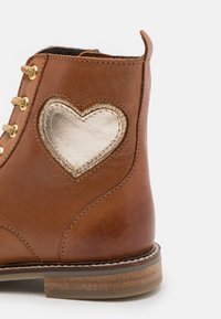 Bota de cuero marrón con ojales dorados, que presenta un acento metálico en forma de corazón dorado en el costado y una suela texturizada y resistente.