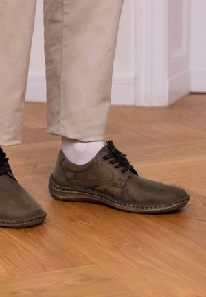 Braune Lederschuhe mit schwarzen Schnürsenkeln, mit runder Spitze und genähten Details. Kombiniert mit beigen Hosen und weißen Socken auf Holzboden.
