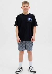 T-shirt nera con grafica a onde e testo, abbinata a pantaloni corti blu a fantasia. Calzini bianchi e sneaker neri completano l'outfit.