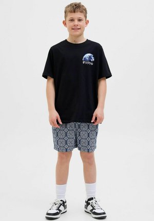 T-shirt noir avec un graphique de vagues et du texte, associé à un short bleu à motifs. Des chaussettes blanches et des baskets noires complètent la tenue.
