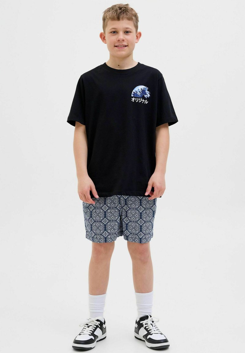T-shirt nera con grafica a onde e testo, abbinata a pantaloni corti blu a fantasia. Calzini bianchi e sneaker neri completano l'outfit.