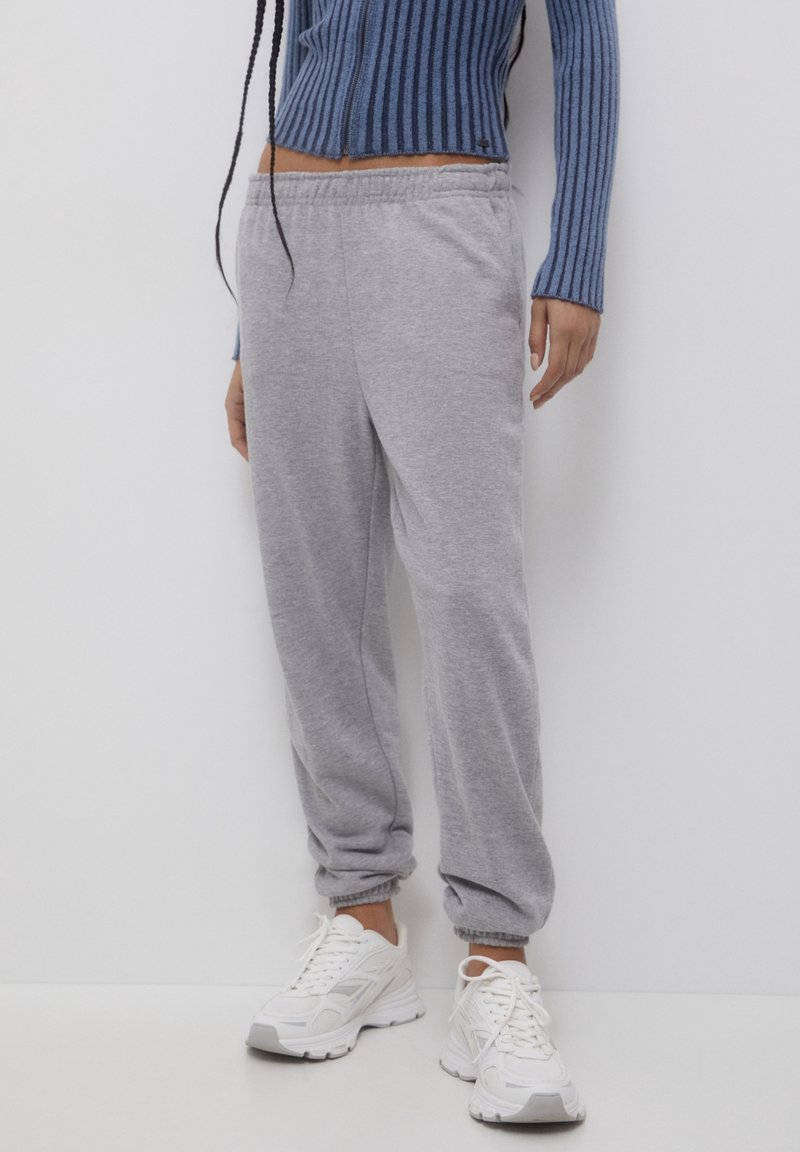Personne portant un pantalon de jogging gris à taille élastique, des baskets blanches et un haut court bleu côtelé à manches longues, debout devant un fond blanc.
