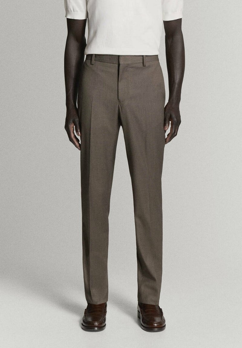 Homme portant un pantalon marron sur mesure, une chemise blanche à manches courtes et des chaussures habillées marron foncé, debout devant un fond uni.