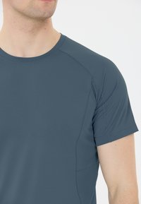 Harmaa lyhythihainen urheilupaita, joka on valmistettu teksturoidusta kankaasta. Siinä on pyöreä kaula-auko ja sauman yksityiskohtia harteilla, mikä luo istuvan ilmeen.