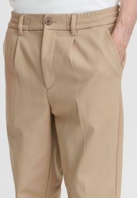 Beige tailored trousers with a smooth texture, featuring a button closure, side pockets, and a slightly tapered design.