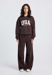 Brown cotton sweatshirt with “USA” and “New York City” printed in white and maroon, paired with matching wide-leg pants and white sneakers.