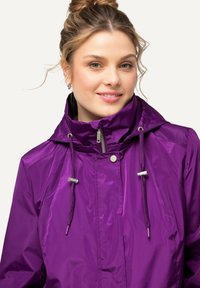 Lila vindjacka med hög krage, justerbara snörningar och silverdetaljer. Slät, lätt tygyta.