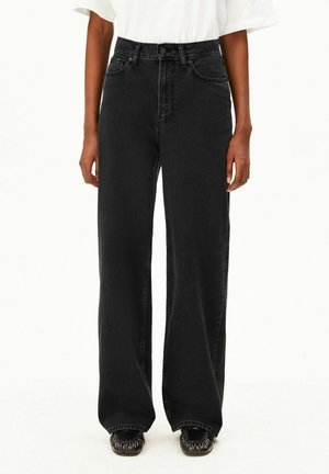 Personne portant un jean noir taille haute à jambes larges, un haut blanc à manches courtes et des chaussures noires à bout froncé, debout devant un fond blanc.