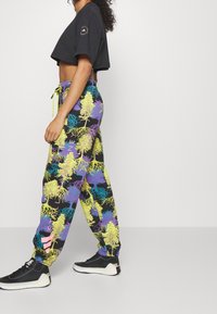 Woman walking wearing black cropped t-shirt and colorful tree-patterned joggers with black and white sneakers on plain background.