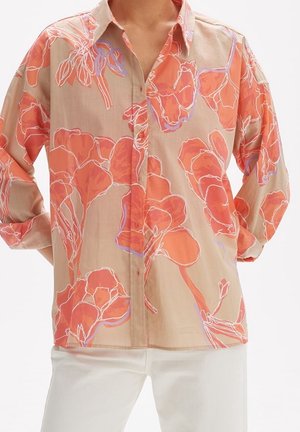 Chemise beige boutonnée avec un large imprimé floral orange portée avec un pantalon blanc, manches retroussées, mains sur les hanches.