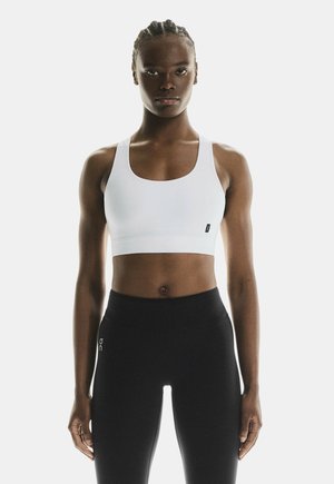 Atletisch persoon met gevlochten haar, gekleed in een witte sport-bh en zwarte legging tegen een effen achtergrond.