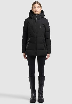 Femme debout portant un manteau matelassé noir à capuche, des leggings noirs et des bottes noires montantes sur un fond clair uni.