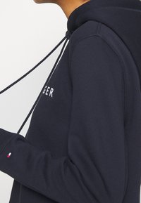 Personne portant un sweat à capuche bleu marine avec des cordons, logo visible sur la manche et texte partiel en blanc sur la poitrine, le tout sur un fond uni.