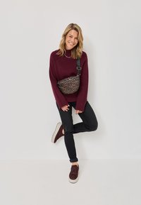 Sacoche en imprimé léopard avec une bandoulière noire, pull bordeaux, jean noir et baskets bordeaux à motifs sur un fond neutre.