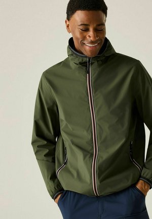 Mann lächelt, trägt eine olivgrüne Jacke mit Reißverschluss und Kapuze und hat die Hände in den Taschen einer marineblauen Hose vor einem einfarbigen Hintergrund.