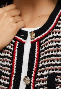 Gestrickte Strickjacke mit einem gestreiften Muster in Schwarz, Rot und Weiß. Verfügt über goldene Knöpfe und einen Rundhalsausschnitt. Texturierte Stoffdetails sind sichtbar.