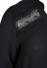 Blusa nera con un pannello in pizzo trasparente sulla parte superiore del petto con motivi floreali, tessuto texturizzato e una vestibilità rilassata.