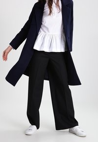 Cappotto lungo blu navy sopra una maglietta peplum bianca, abbinato a pantaloni neri a gamba larga e sneakers bianche. Le caratteristiche includono texture e colori a contrasto.