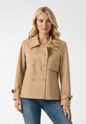 Donna con capelli biondi che indossa una giacca doppiopetto beige con dettagli a fiocco sulle maniche e jeans blu, in piedi davanti a uno sfondo semplice.