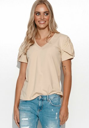 Beżowa koszulka typu V-neck z bufkami na krótkich rękawach; wykonana z miękkiego materiału o gładkiej strukturze; zestawiona z postarzanymi niebieskimi dżinsami.
