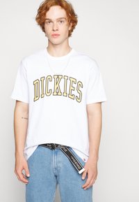 Hvit t-skjorte med korte ermer og fet svart og gull "DICKIES"-trykk. Båret med lyseblå jeans og et svart belte med hvit skrift.