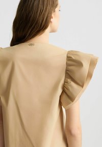 Top beige con maniche a sbuffo, una texture liscia e una chiusura decorativa in oro sul collo posteriore. Design semplice ed elegante.