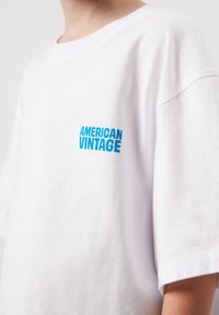 Biele tričko nosené osobou, s malým modrým nápisom "AMERICAN VINTAGE" na ľavej strane hrudníka.