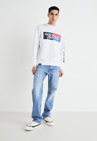 Sudadera gris clara con un audaz logo en negro y rojo de "Red Bull Racing". Combinada con pantalones vaqueros azules desgastados y zapatillas blancas con detalles en negro.