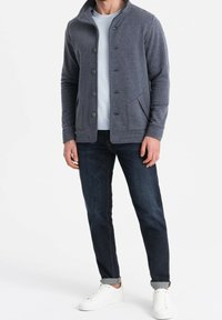 Homme portant une veste grise boutonnée, un t-shirt bleu clair, un jean foncé et des baskets blanches, debout sur un fond blanc uni.