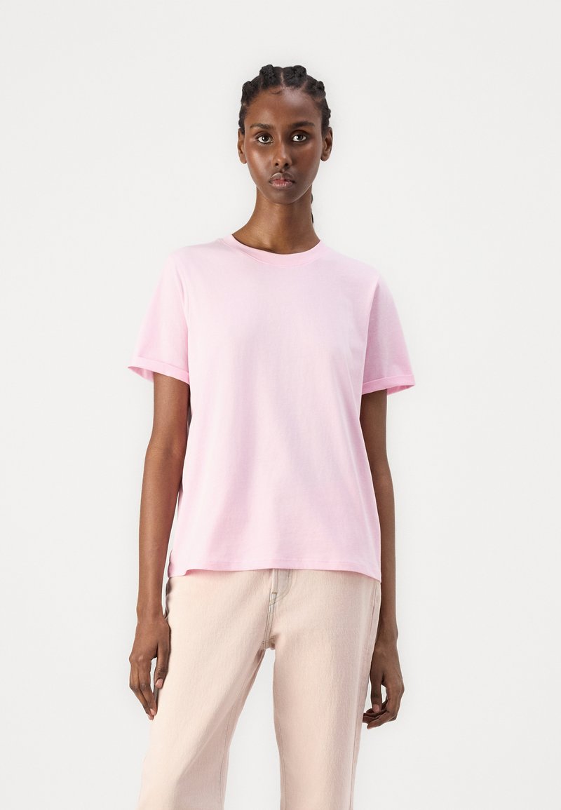 Lys rosa bomull t-skjorte med korte ermer og rund hals, brukt sammen med lyse beige bukser. En enkel design, uten mønstre eller aksenter.