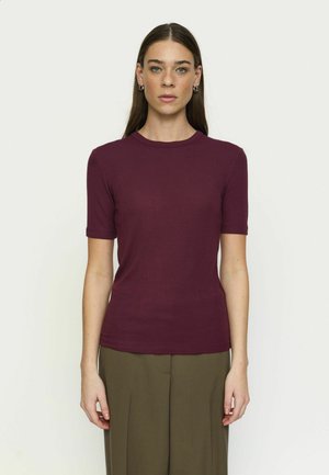 Donna con lunghi capelli castani indossa una maglietta bordeaux aderente a maniche corte e pantaloni larghi verde oliva, in piedi su uno sfondo bianco semplice.