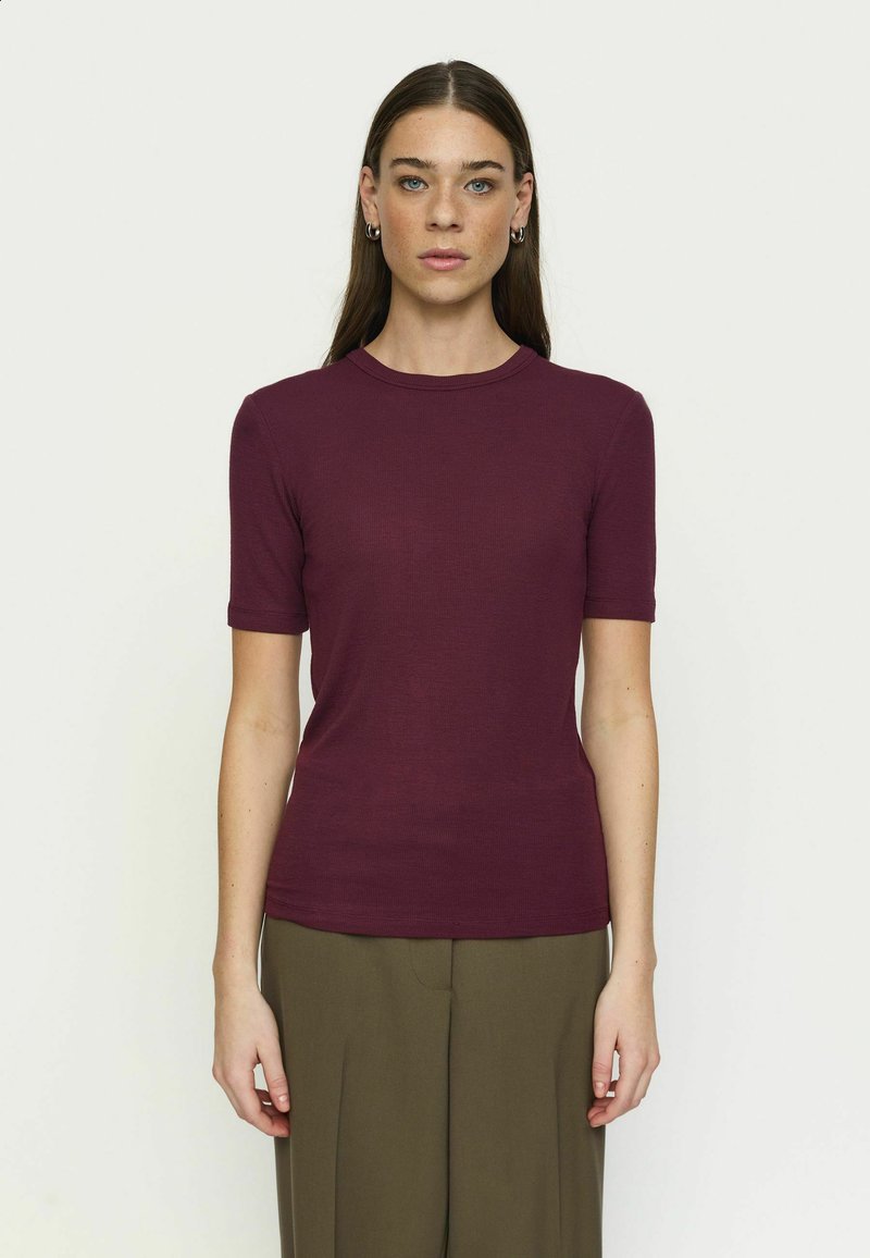 Femme aux longs cheveux bruns portant un haut bordeaux ajusté à manches courtes et un pantalon ample vert olive, debout sur un fond blanc uni.