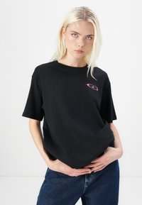 T-shirt en coton noir à manches courtes, avec un petit graphisme rose sur la poitrine. Coupe décontractée, design minimaliste, texture douce.