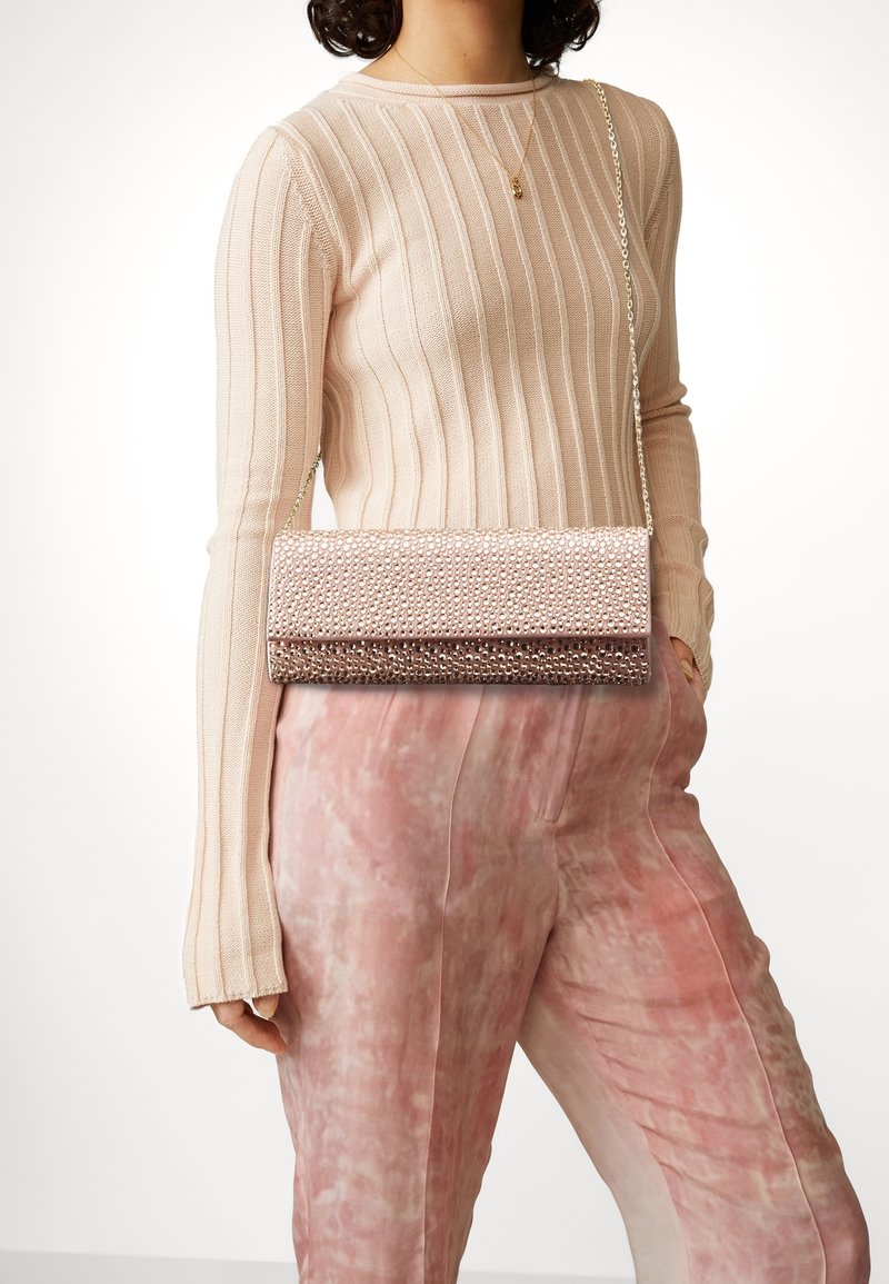 Femme portant un pull beige côtelé à manches longues, un pantalon tie-dye rose et un sac bandoulière rose clouté.