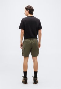 Άνδρας με την πλάτη γυρισμένη φοράει μαύρο μπλουζάκι, χακί cargo σορτς με πλαϊνές τσέπες, μαύρες κάλτσες και καφέ ορειβατικά παπούτσια σε λευκό φόντο.